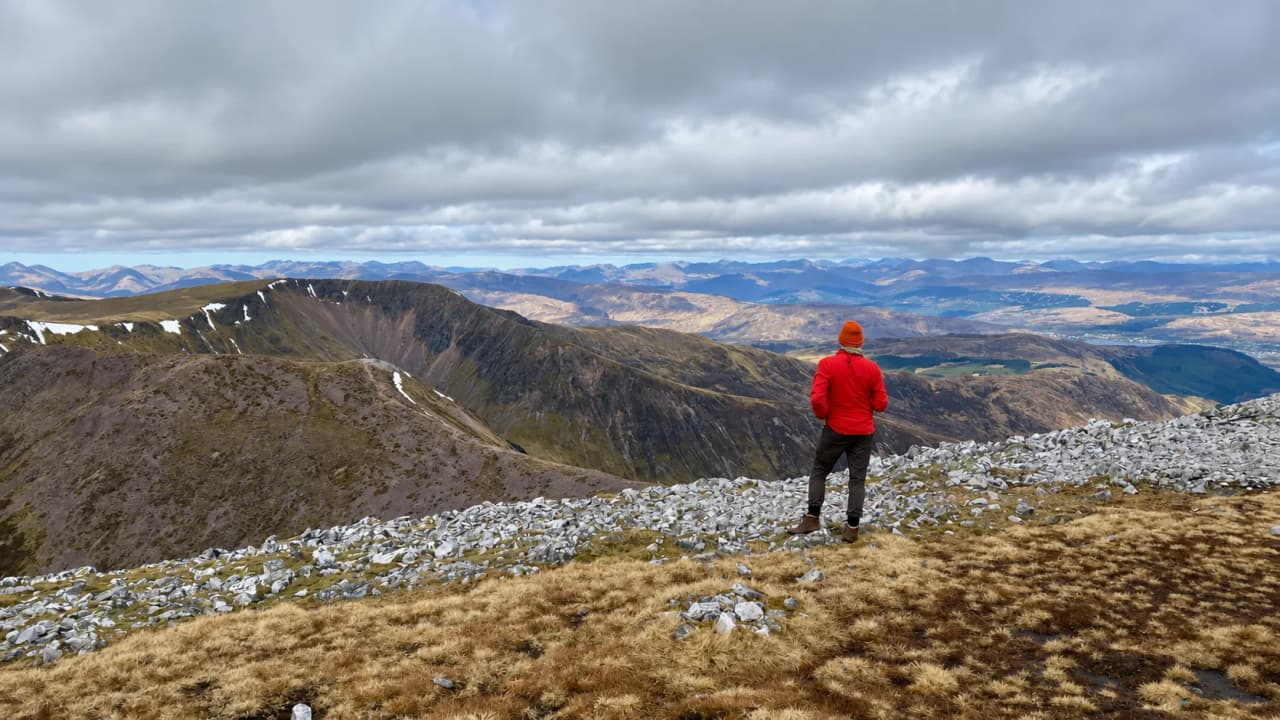 A hiker in a red jacket on a rocky summit, looking out over snow-streaked ridges and Highland glens