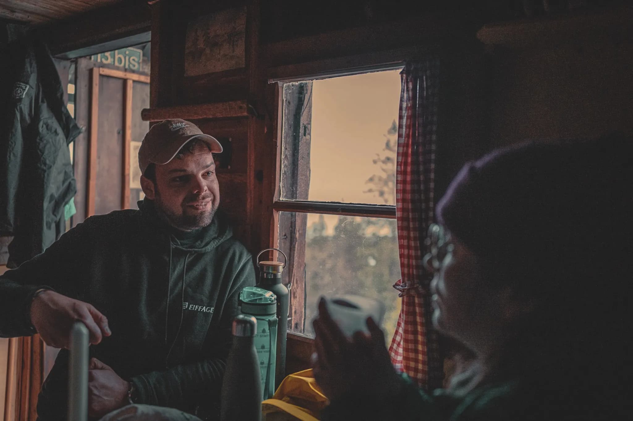 Two people are having a friendly chat inside a wooden chalet, with a view of the countryside.