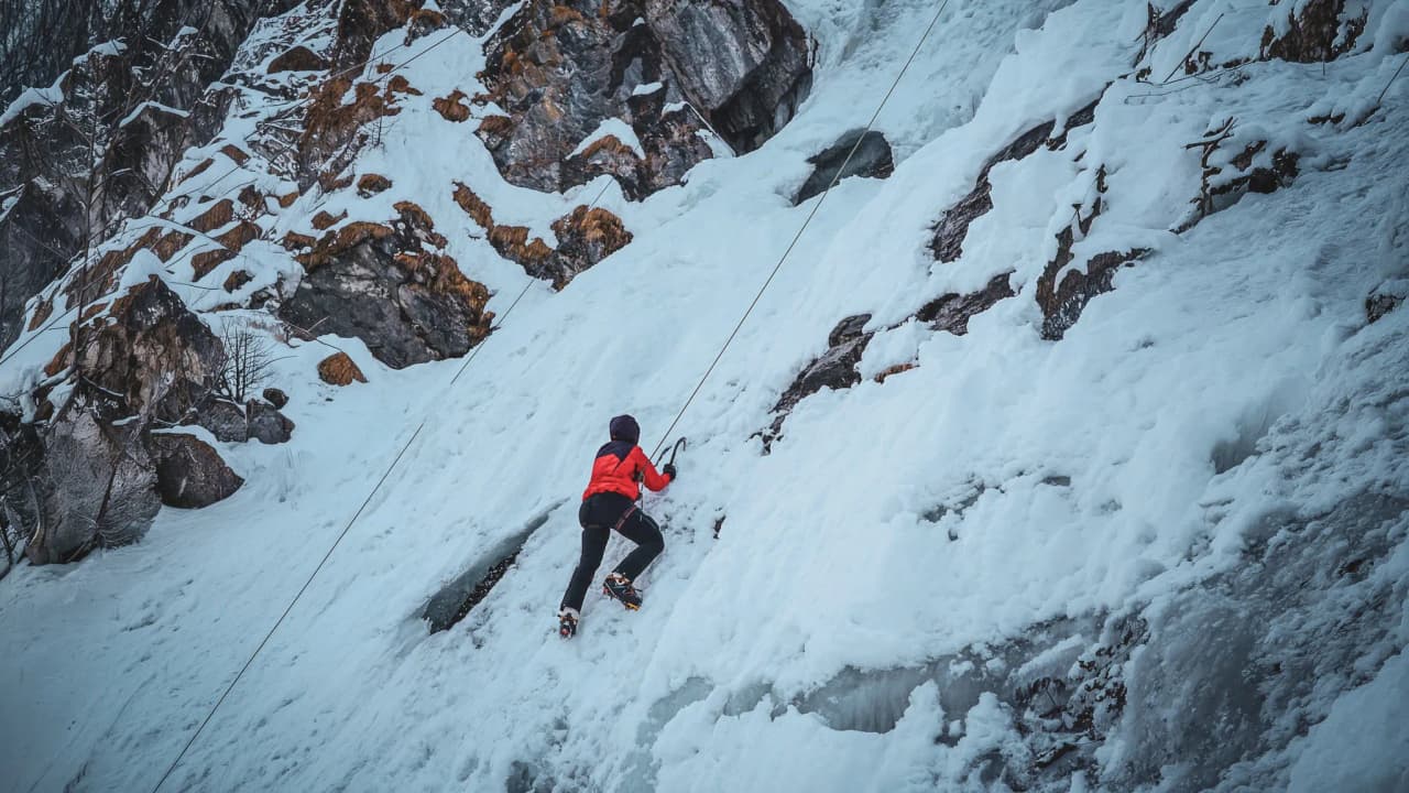 A climber in red climbs an icefall in the Italian Alps under a blue sky.