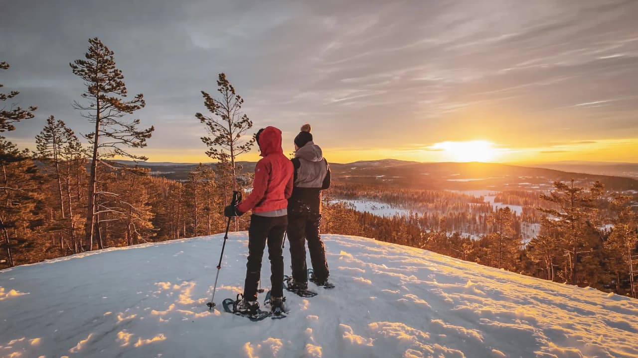 Two hikers watch the sun set over a snow-covered landscape in Swedish Lapland.