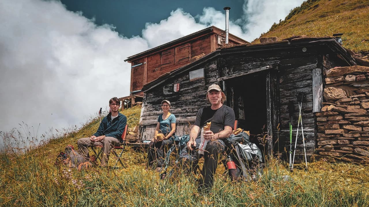 Three hikers relaxing in front of an alpine mountain hut, surrounded by flower-filled meadows and clouds.