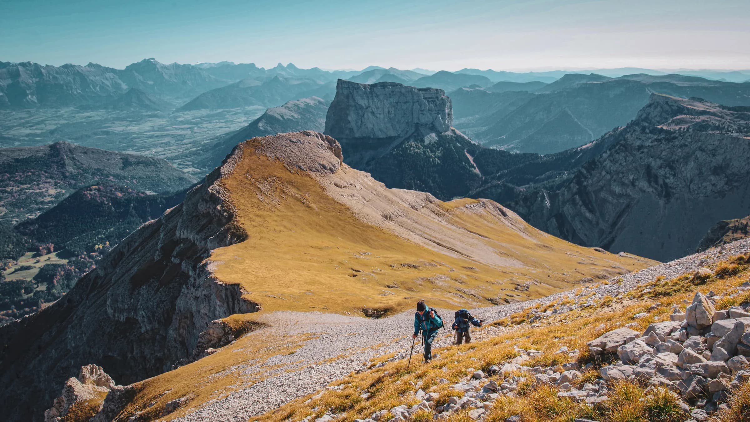 Deux randonneurs gravissent une crête dorée avec des montagnes majestueuses en arrière-plan.