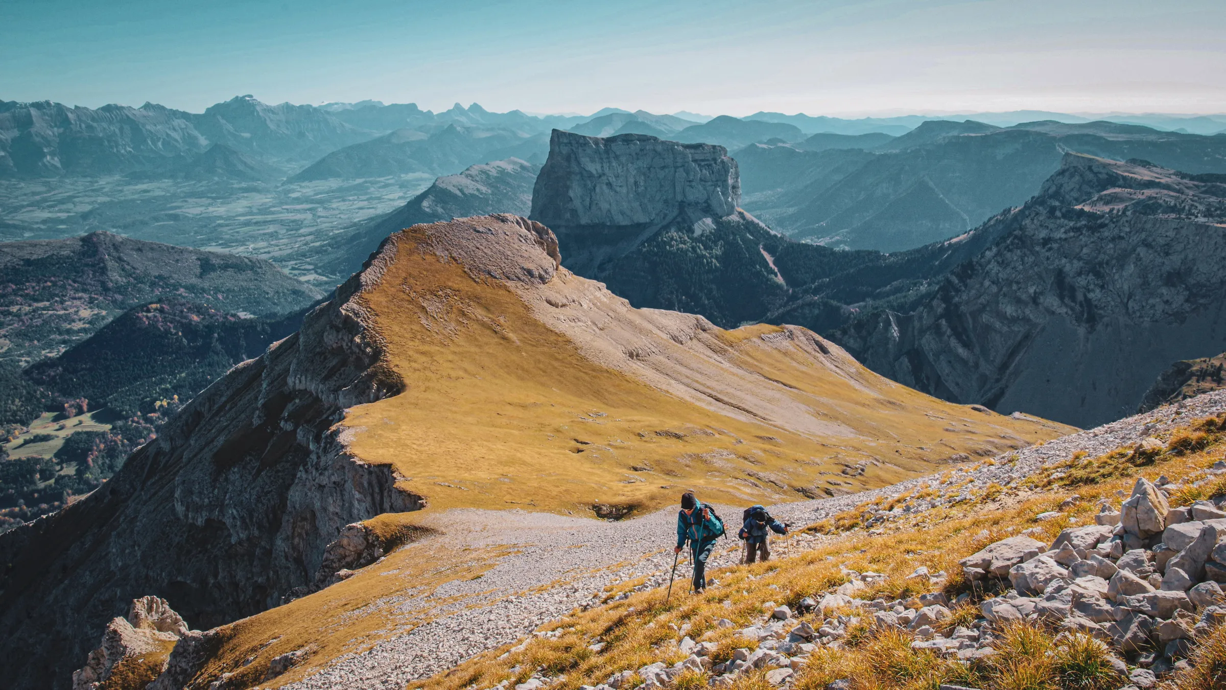 Deux randonneurs gravissent une crête dorée avec des montagnes majestueuses en arrière-plan.