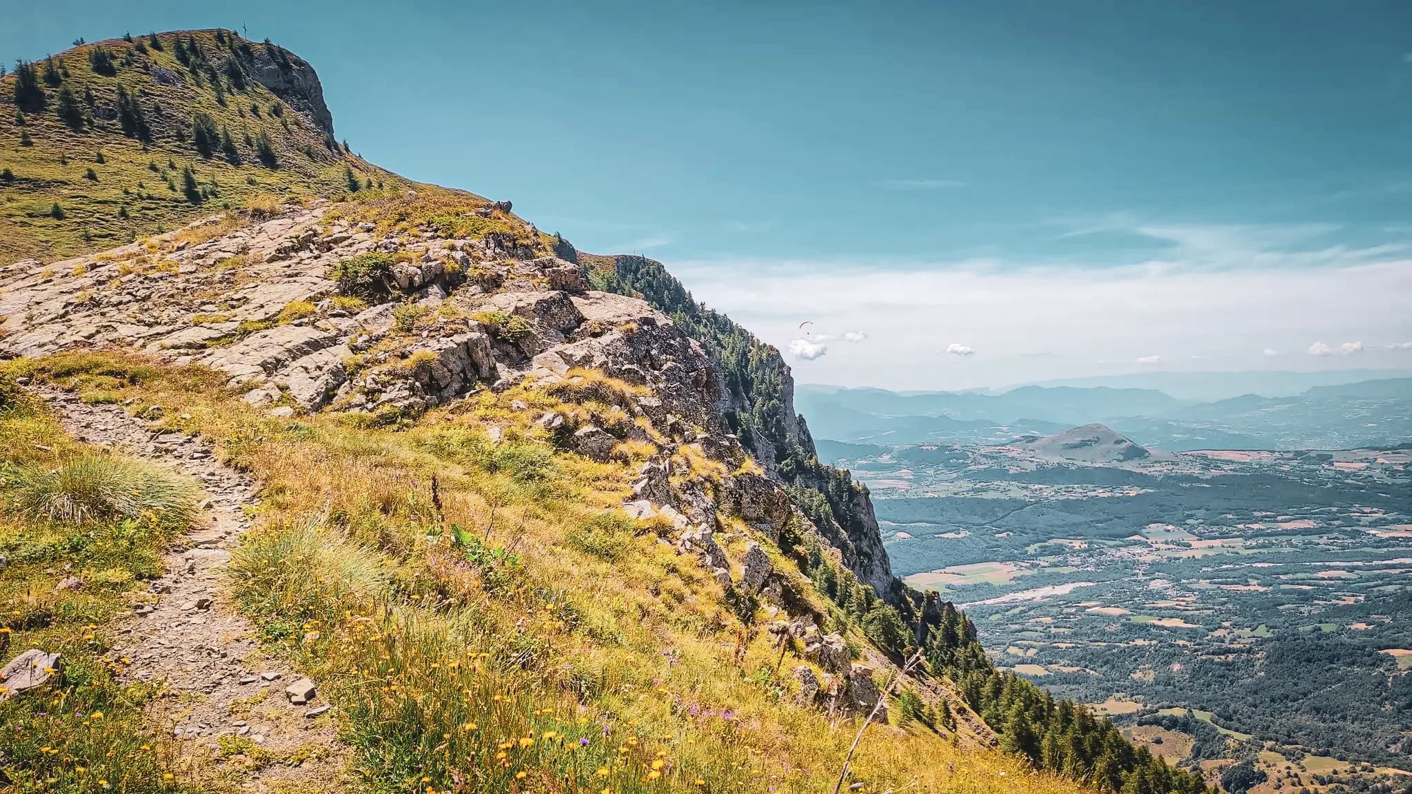 Un chemin sinueux à flanc de montagne, surplombant des vallées verdoyantes sous un ciel azur.