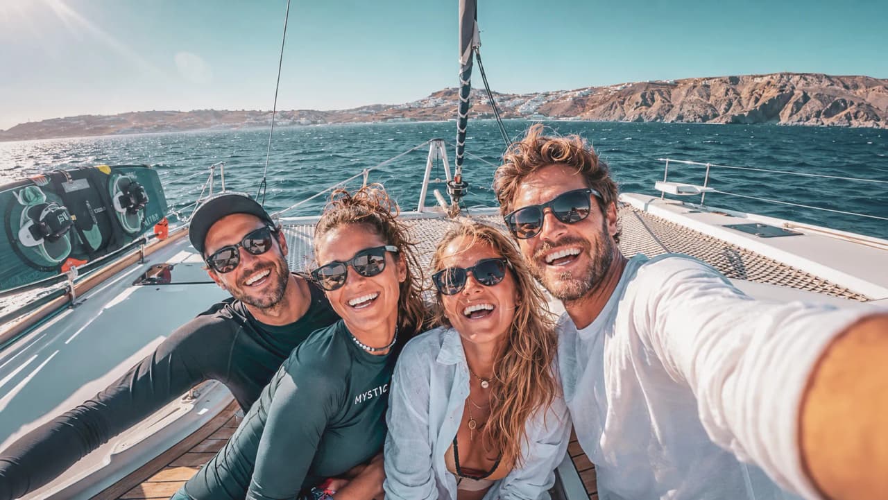 Vier lachende vrienden nemen een selfie op een catamaran, met de zee en eilanden op de achtergrond.