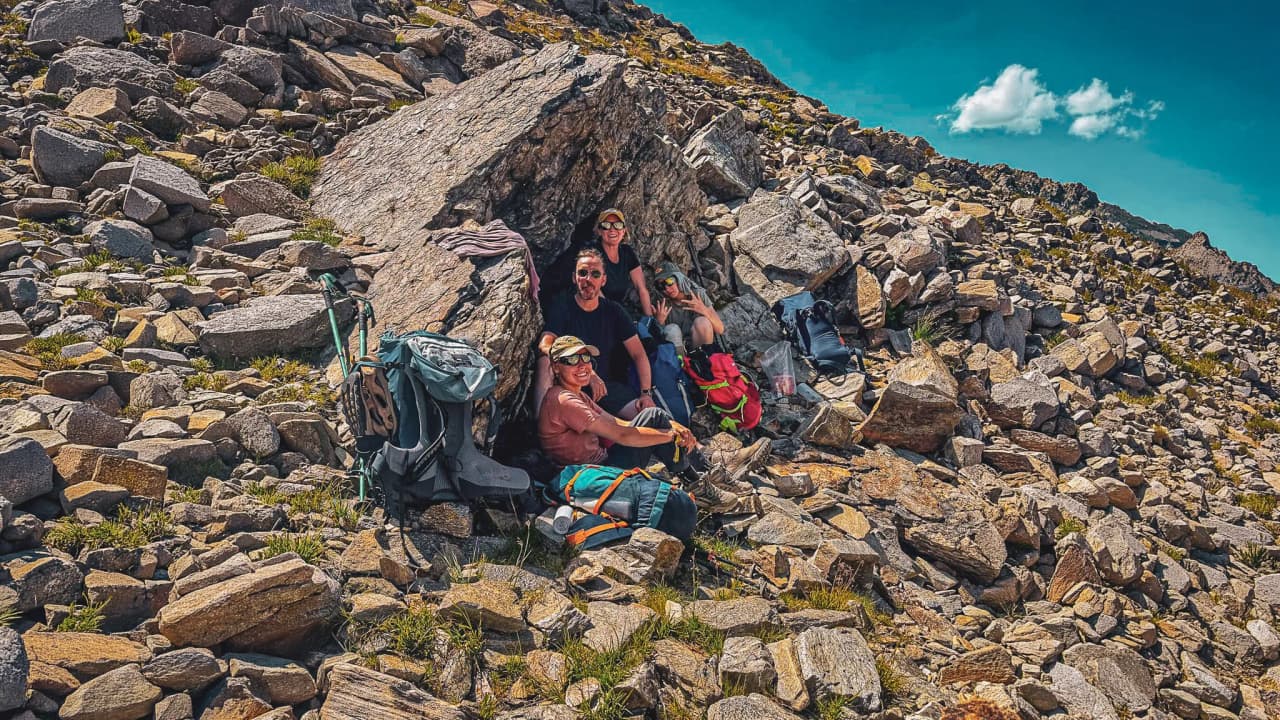 A group of hikers rest under a rock, enjoying the mountain scenery.