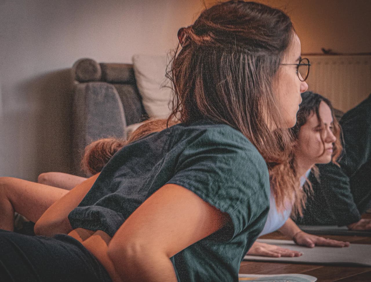 Een groep mensen ligt op hun buik te oefenen in een binnenruimte. Ze lijken zich te concentreren, met matrassen of matten op de grond. Het licht is zacht en warm en creëert een sfeer