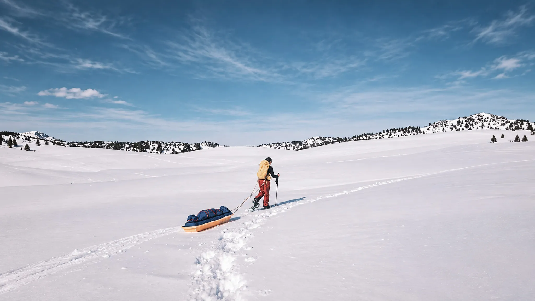 Un aventurier en raquettes tire une pulka à travers un paysage enneigé, sous un ciel bleu.