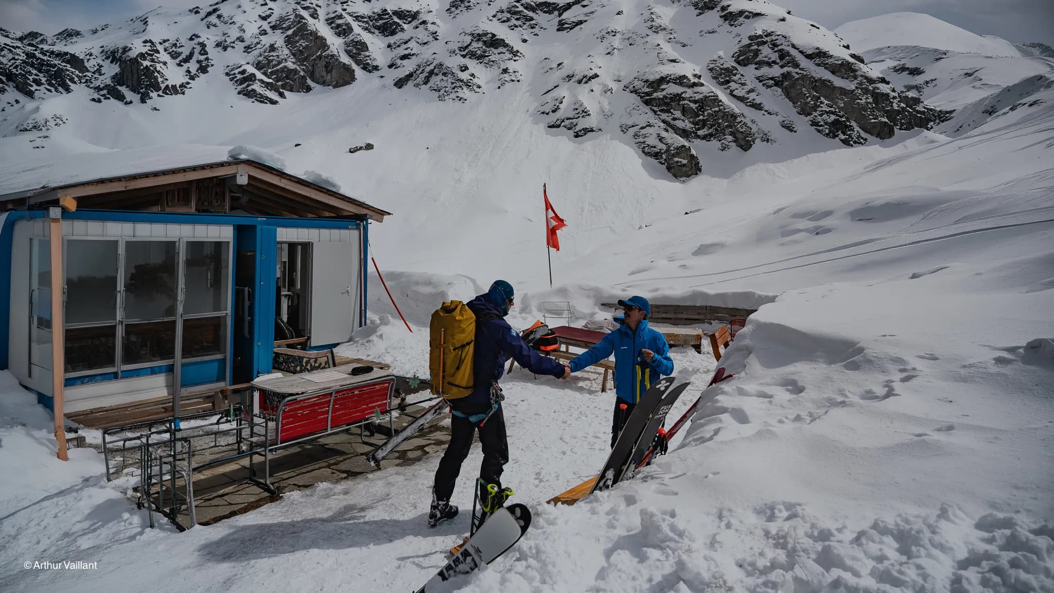 Two skiers greet each other in front of an alpine mountain hut, with snow-capped mountains in the background.