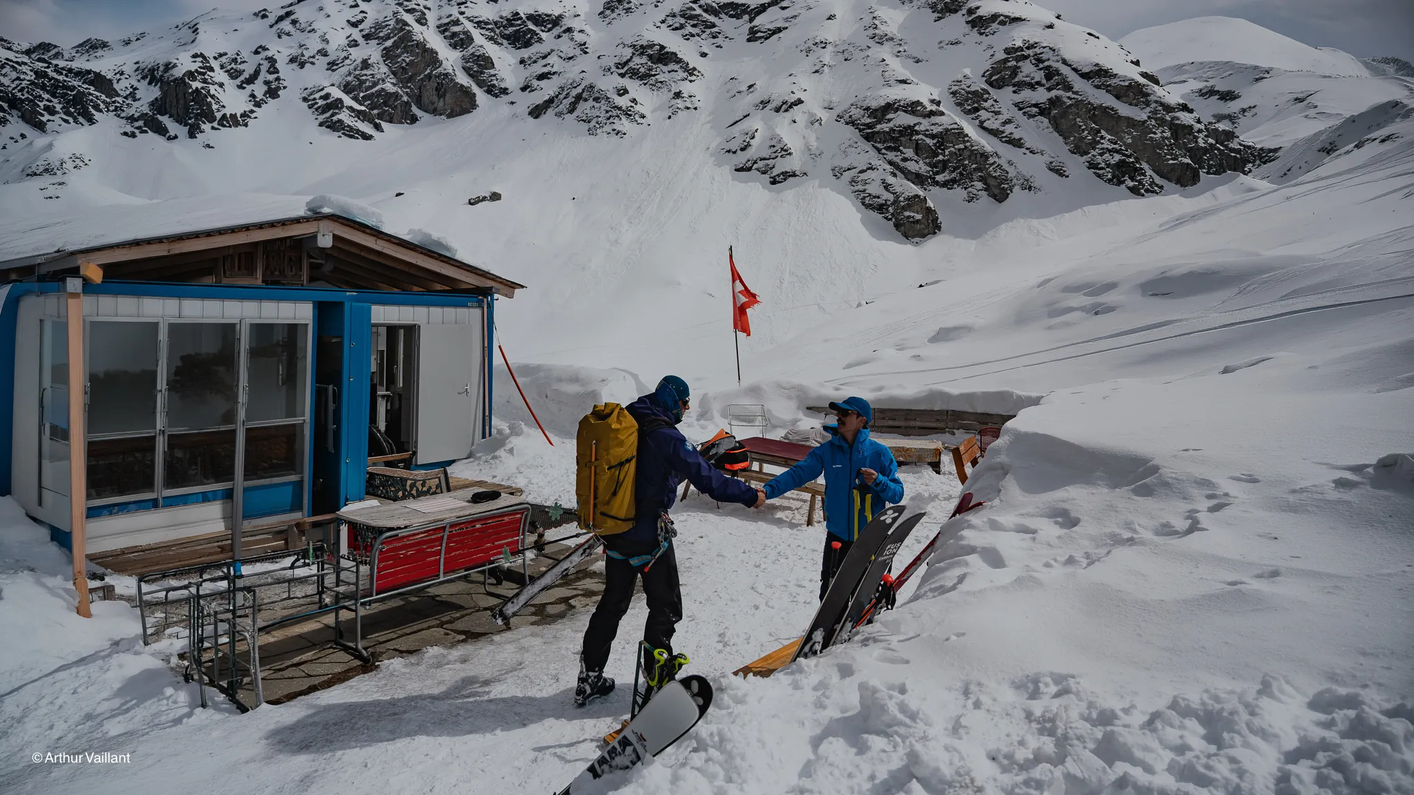 Two skiers greet each other in front of an alpine mountain hut, with snow-capped mountains in the background.