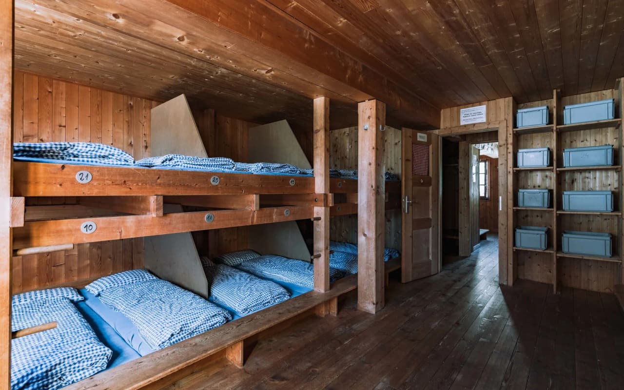 The warm interior of an alpine Mountain hut, with bunk beds and checked bedding, and a friendly atmosphere.