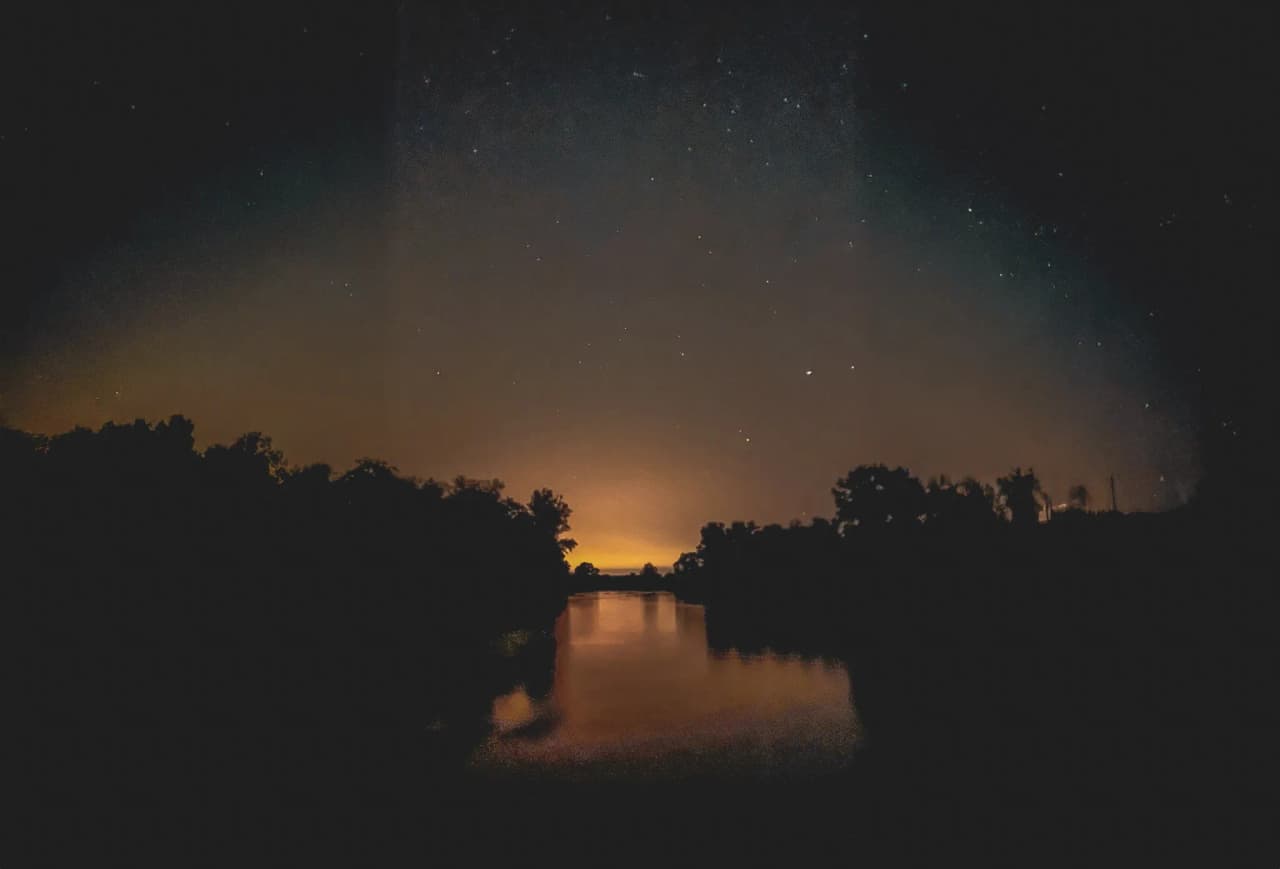 Een diepe sterrenhemel met een vage oranje gloed aan de horizon, die doet denken aan de schemering of stadsverlichting in de verte. Het wateroppervlak is kalm en weerspiegelt de sterren en de silhouetten van donkere bomen.