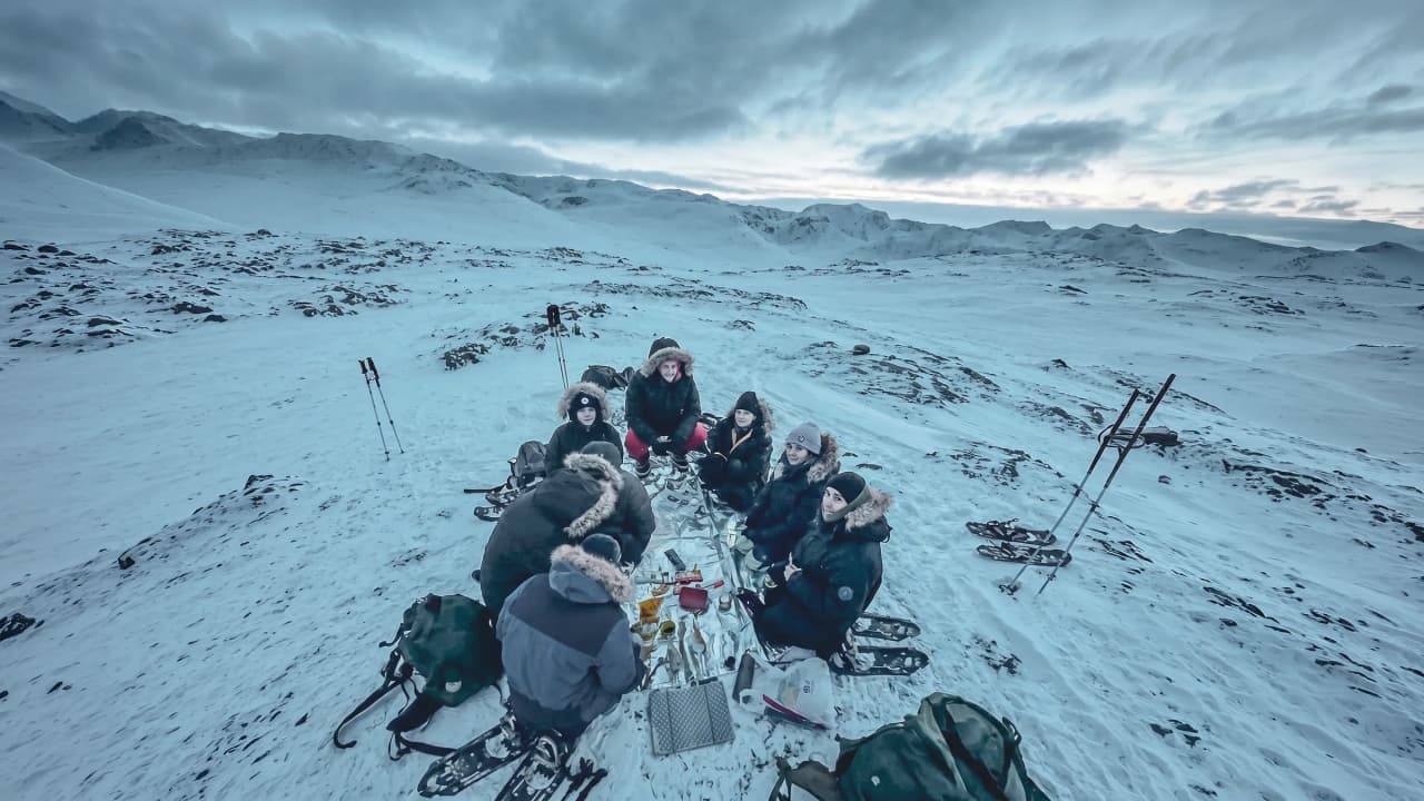A group of adventurers in Lapland, gathered around a table, with snow and mountains in the background.
