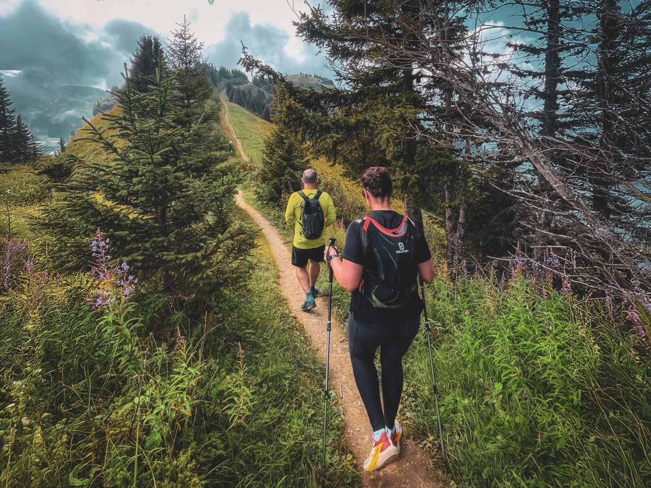 Two hikers follow an alpine path surrounded by greenery, with Mont Blanc in the background.