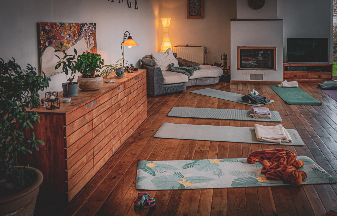 Een warme binnenruimte met een houten vloer. Op de vloer liggen verschillende yogamatten, sommige met opgevouwen handdoeken. Een comfortabele bank, versierd met kussens, staat naast een muur waar je
