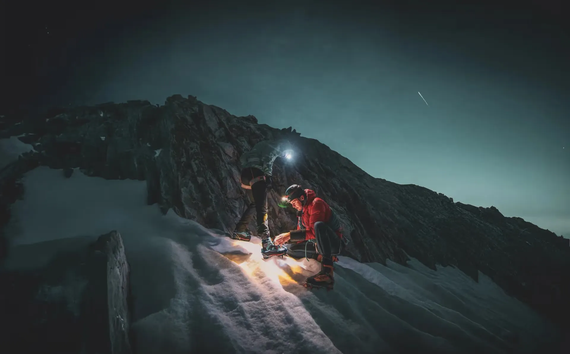 Deux alpinistes en pleine ascension nocturne sur la neige éclairée par des lampes frontales, avec des montagnes majestueuses en arrière-plan.