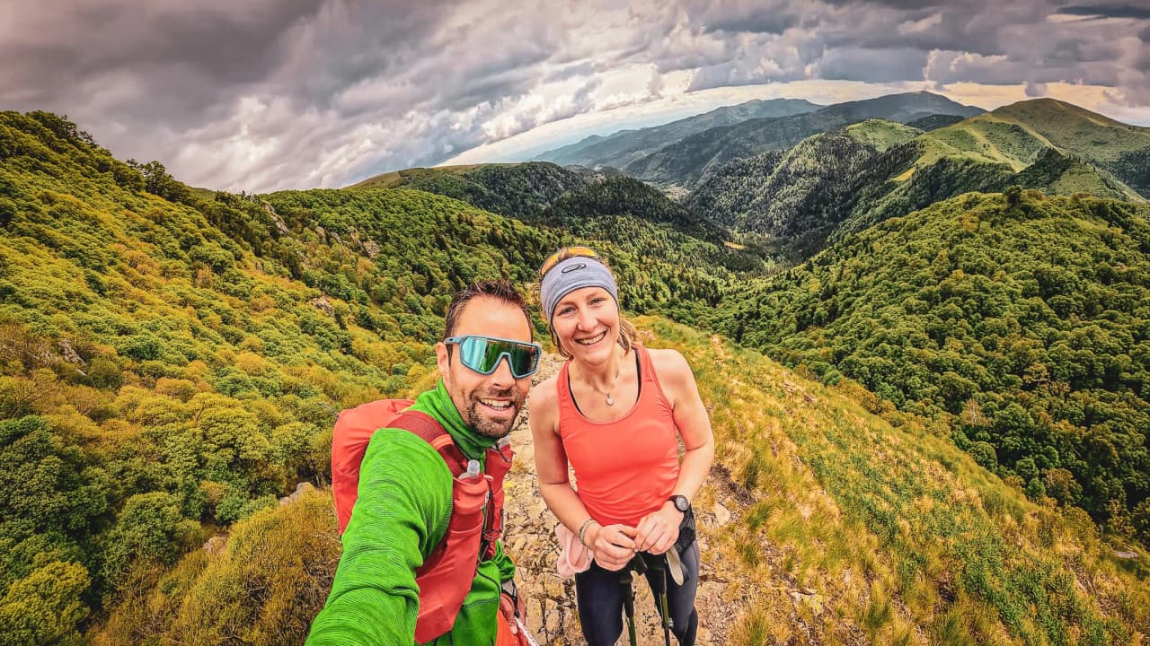 Deux randonneurs souriants prennent un selfie avec des montagnes verdoyantes en arrière-plan.