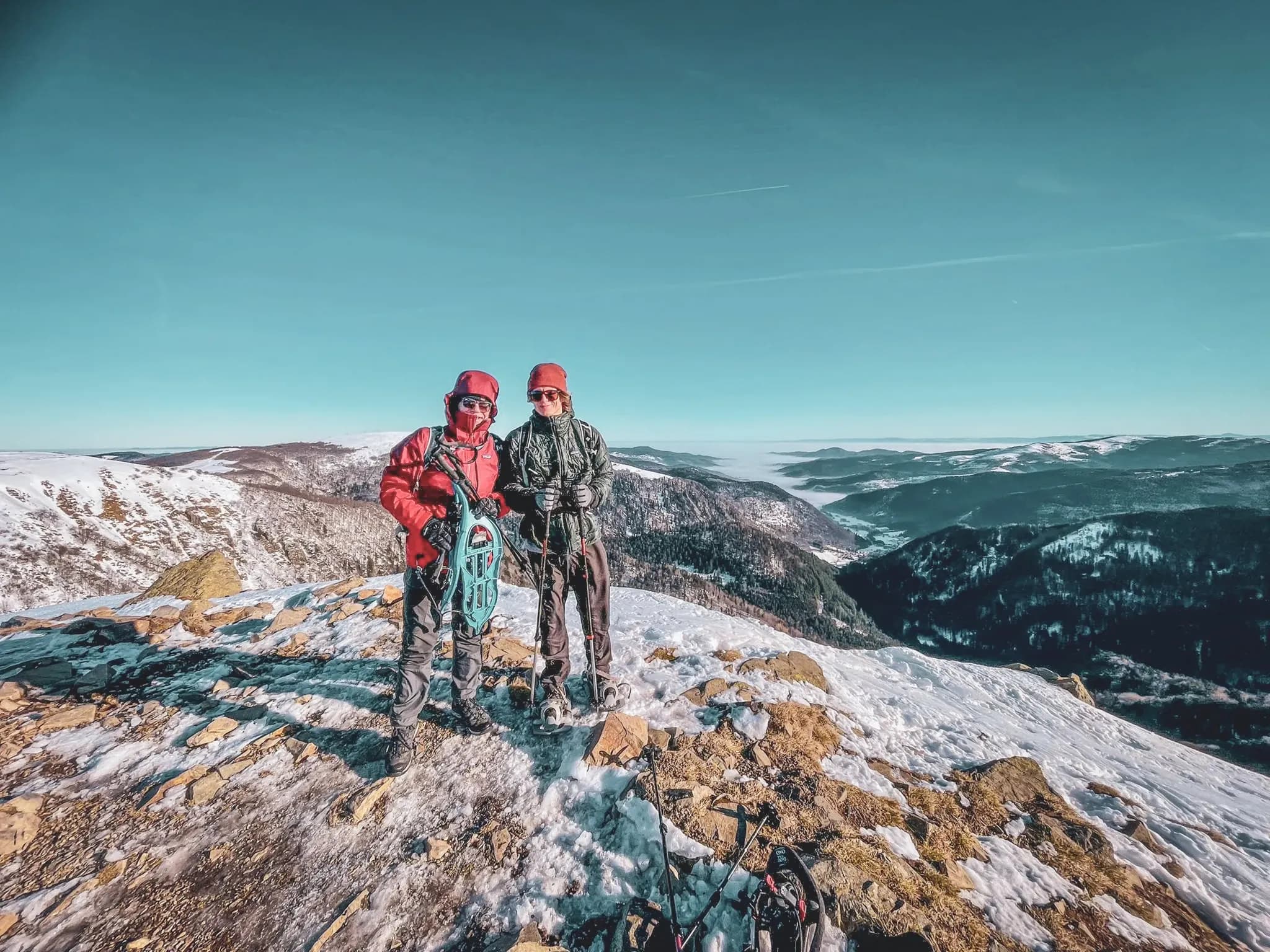 Two hikers on snowshoes, at the top of the Vosges mountains, surrounded by snow and mountain scenery.