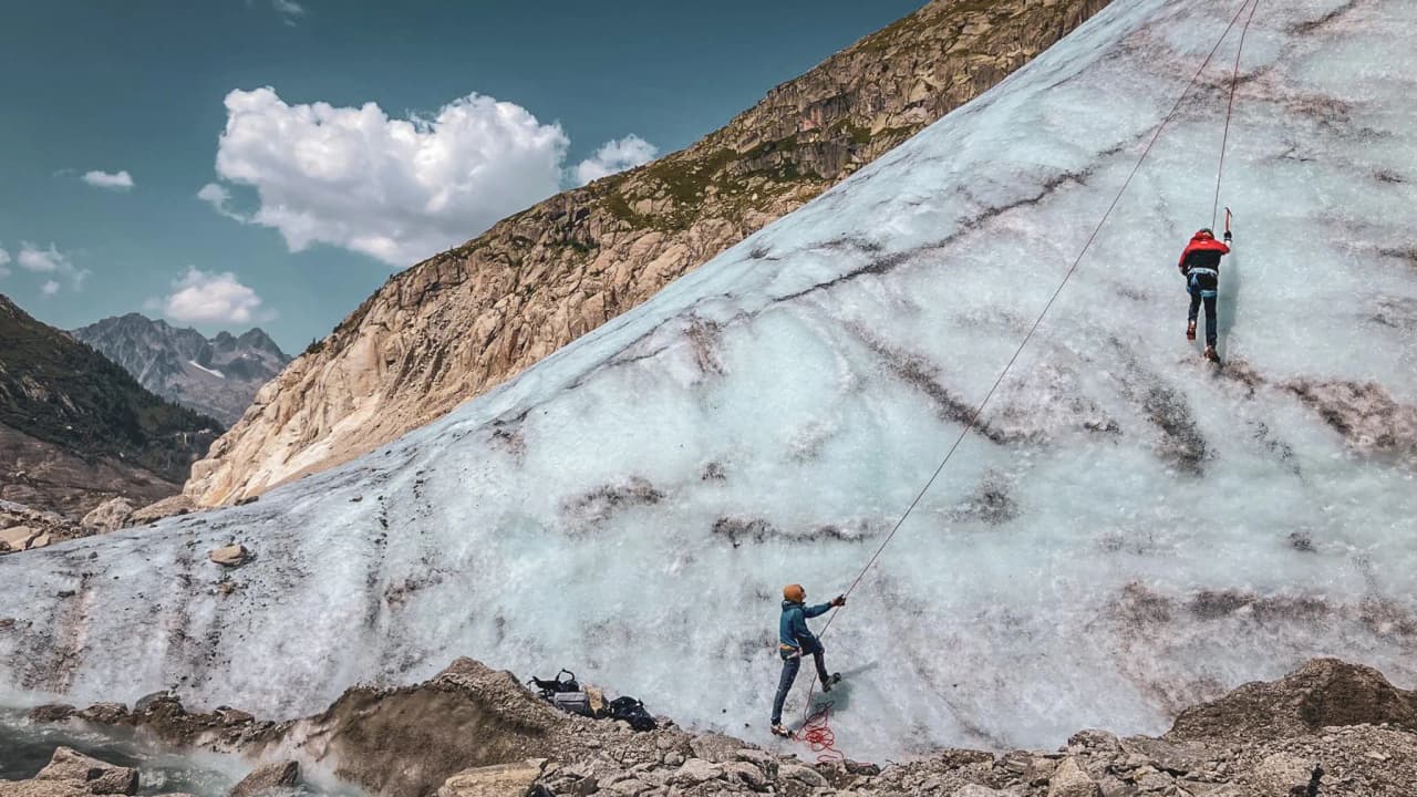Deux alpinistes escaladent un glacier majestueux sous un ciel bleu, entourés de montagnes.