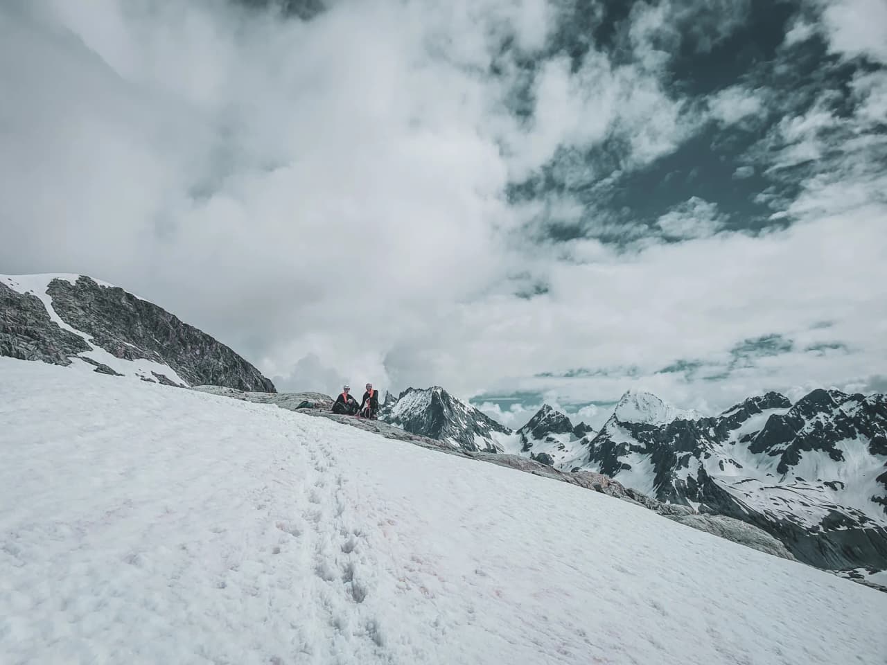 Two adventurers rest on a snowy ridge, surrounded by majestic Alpine peaks.