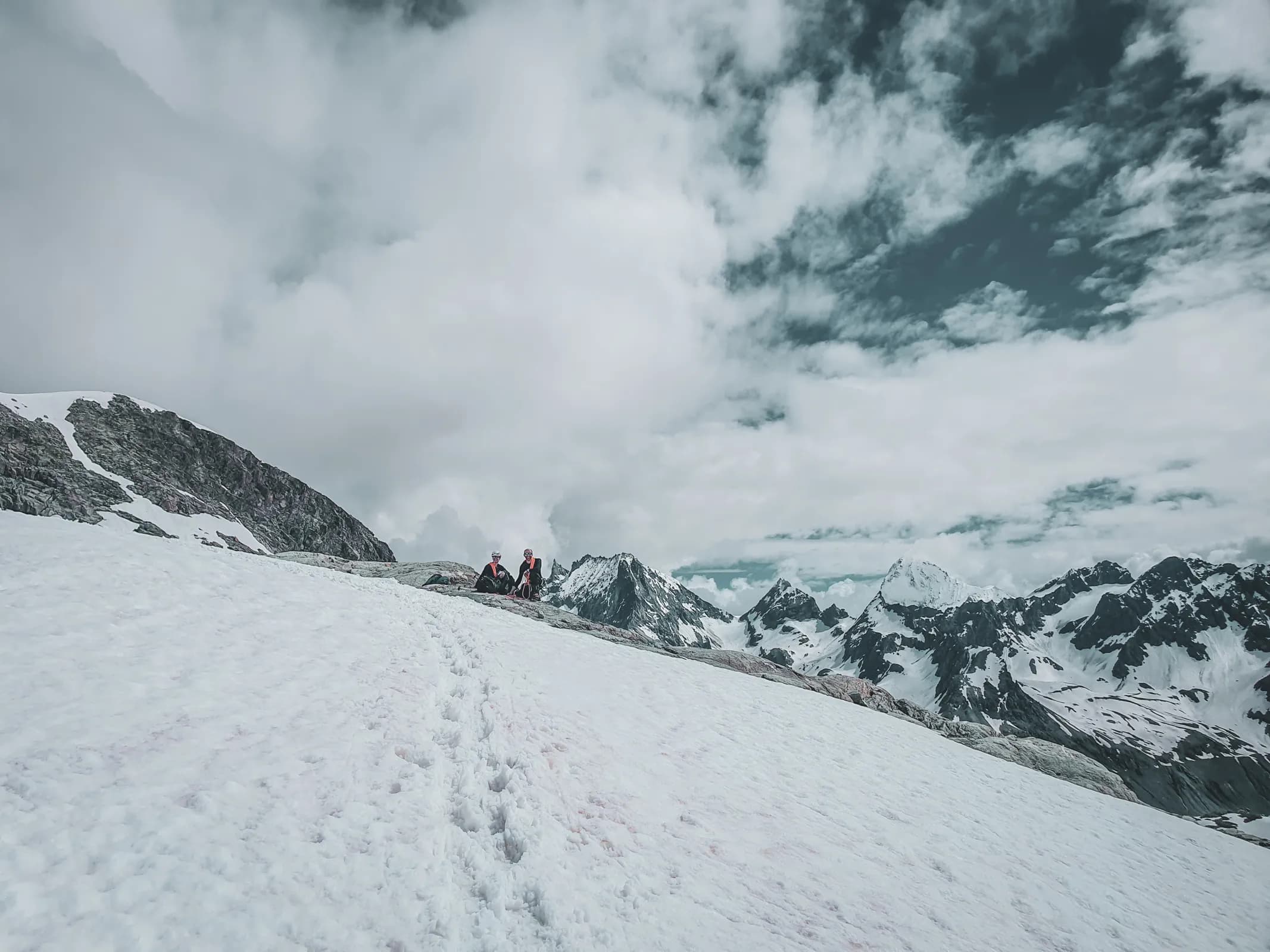 Two adventurers rest on a snowy ridge, surrounded by majestic Alpine peaks.