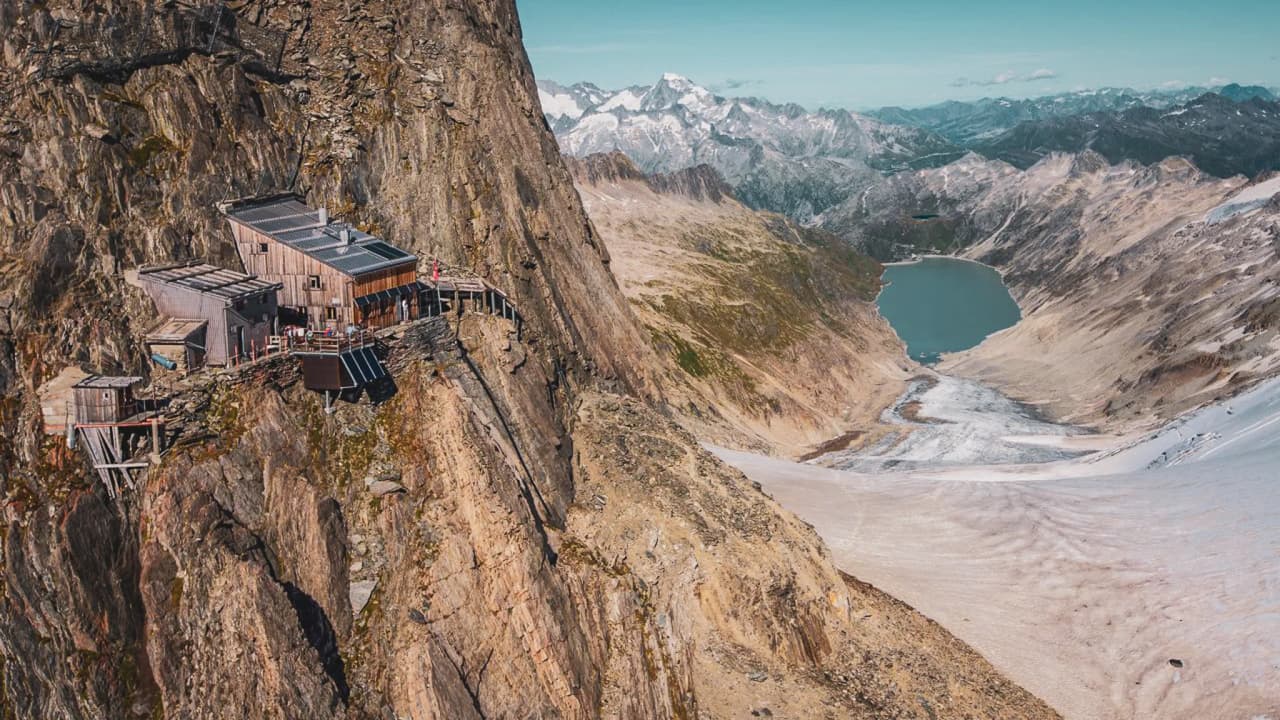 Refuge en bois perché sur un rocher, entouré de montagnes majestueuses et d'un glacier.