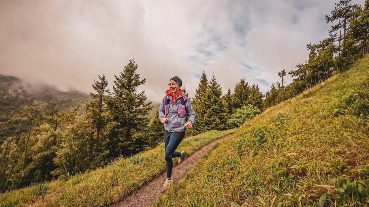 Une joggeuse souriante court sur un sentier verdoyant des Vosges, entourée de nature.