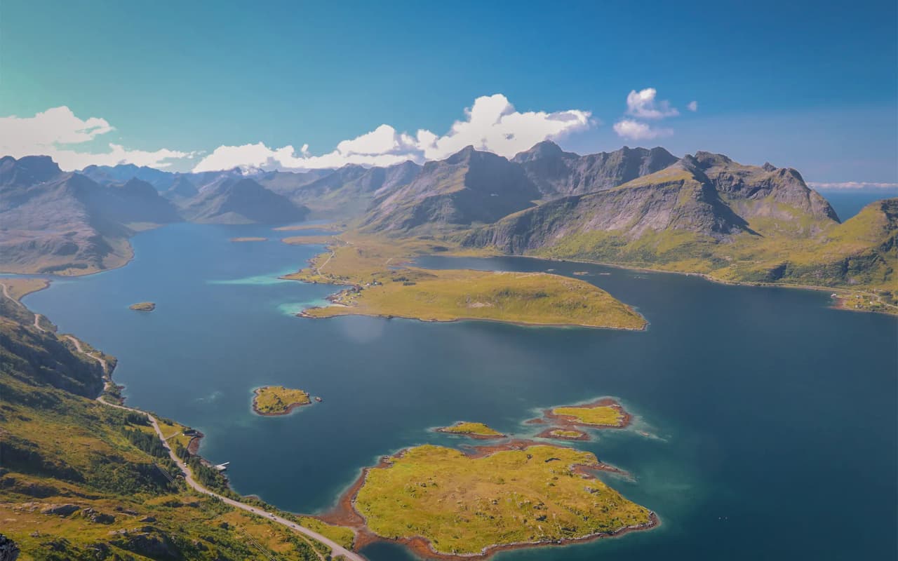 A majestic mountain landscape with a large expanse of water in the foreground, reflecting the surrounding mountains. Green hills and trees line the shore, while several houses, including one with a