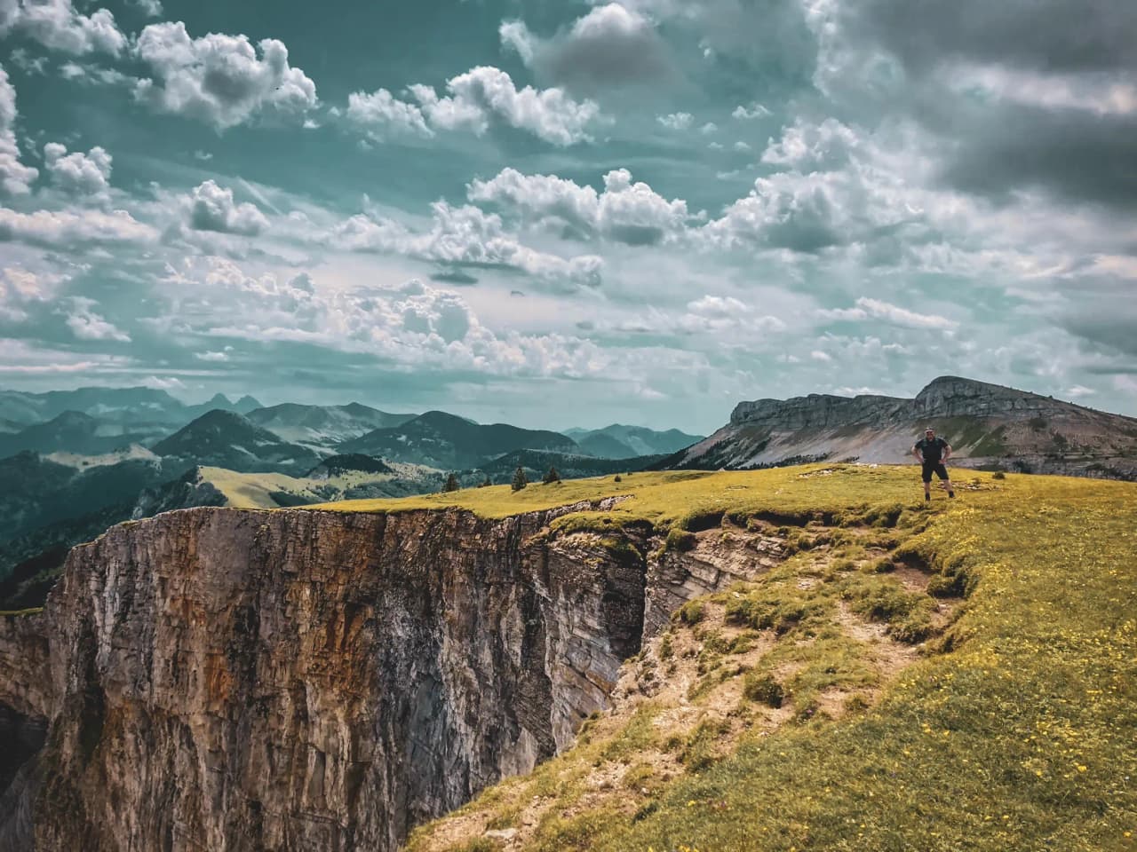 A hiker overlooks green ridges and majestic mountains under a cloudy sky.