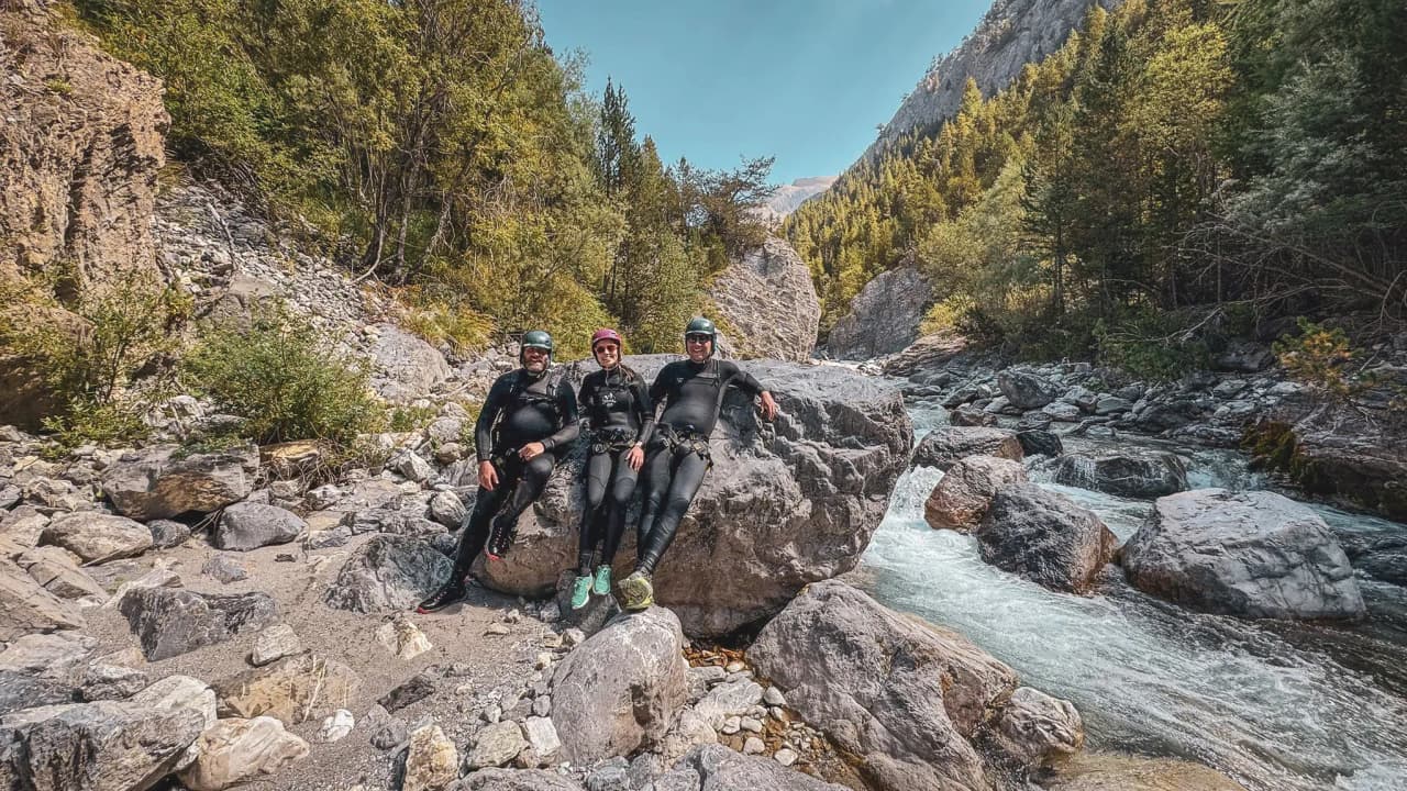 Three adventurers in wetsuits, posing on a rock beside a swift-flowing river in an Alpine setting.