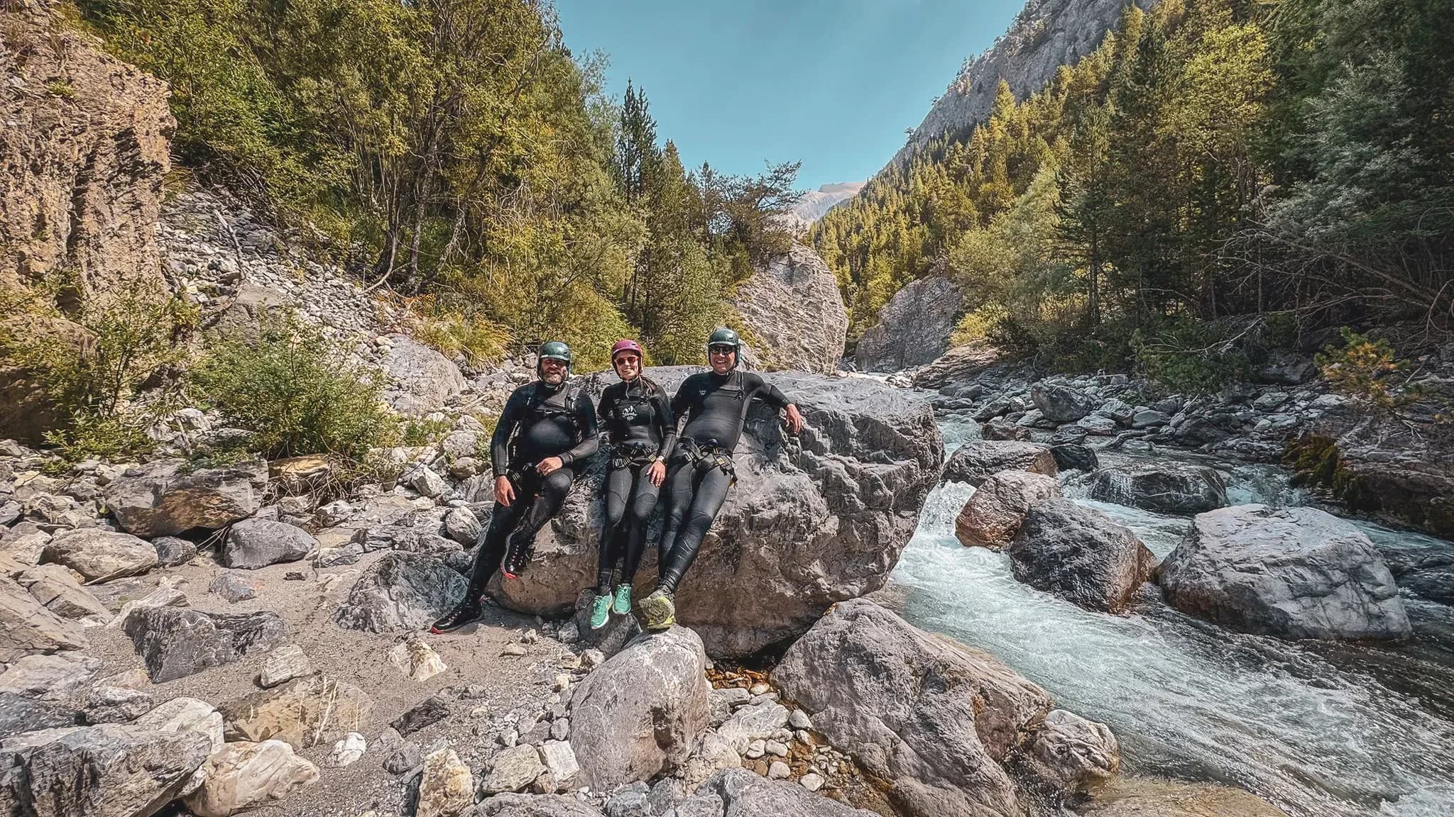 Three adventurers in wetsuits, posing on a rock beside a swift-flowing river in an Alpine setting.