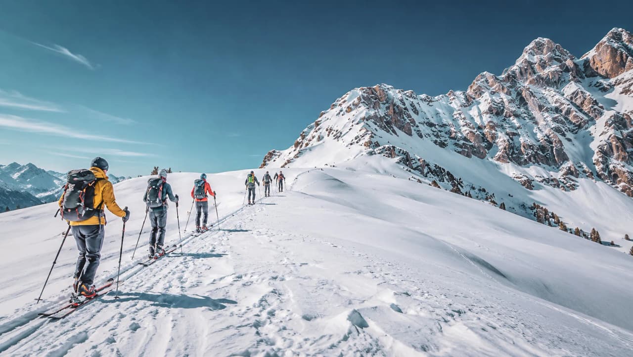 A group of ski tourers crossing a snow-covered alpine landscape beneath a brilliant blue sky.
