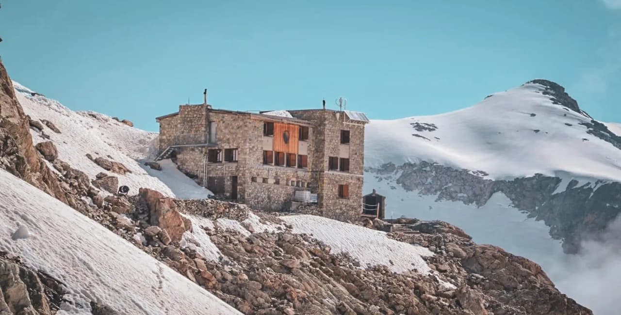 Un refuge en pierre sur une crête alpine, entouré de neige et de montagnes majestueuses.