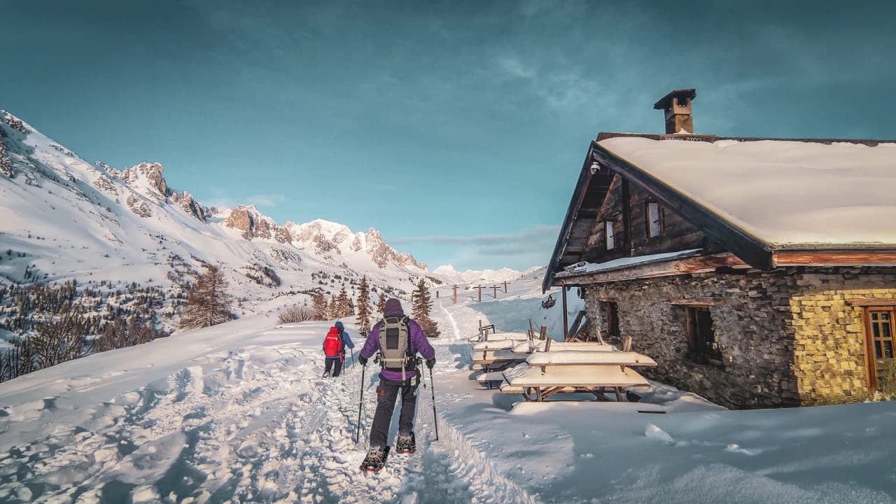 Snowshoe hikers in front of a picturesque mountain hut set against a majestic snow-covered landscape.