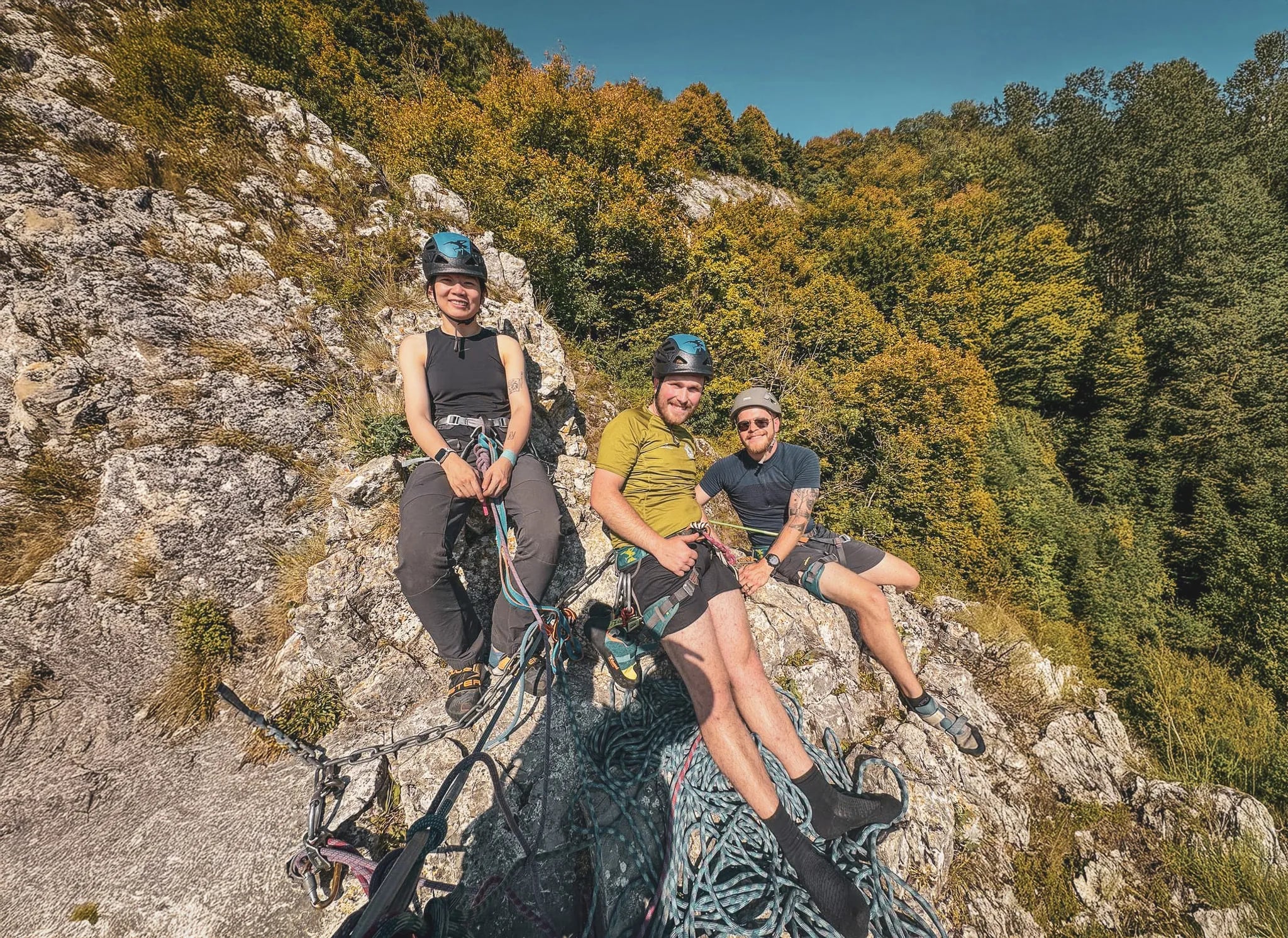 Three smiling climbers rest on a sunny cliff, surrounded by lush nature.