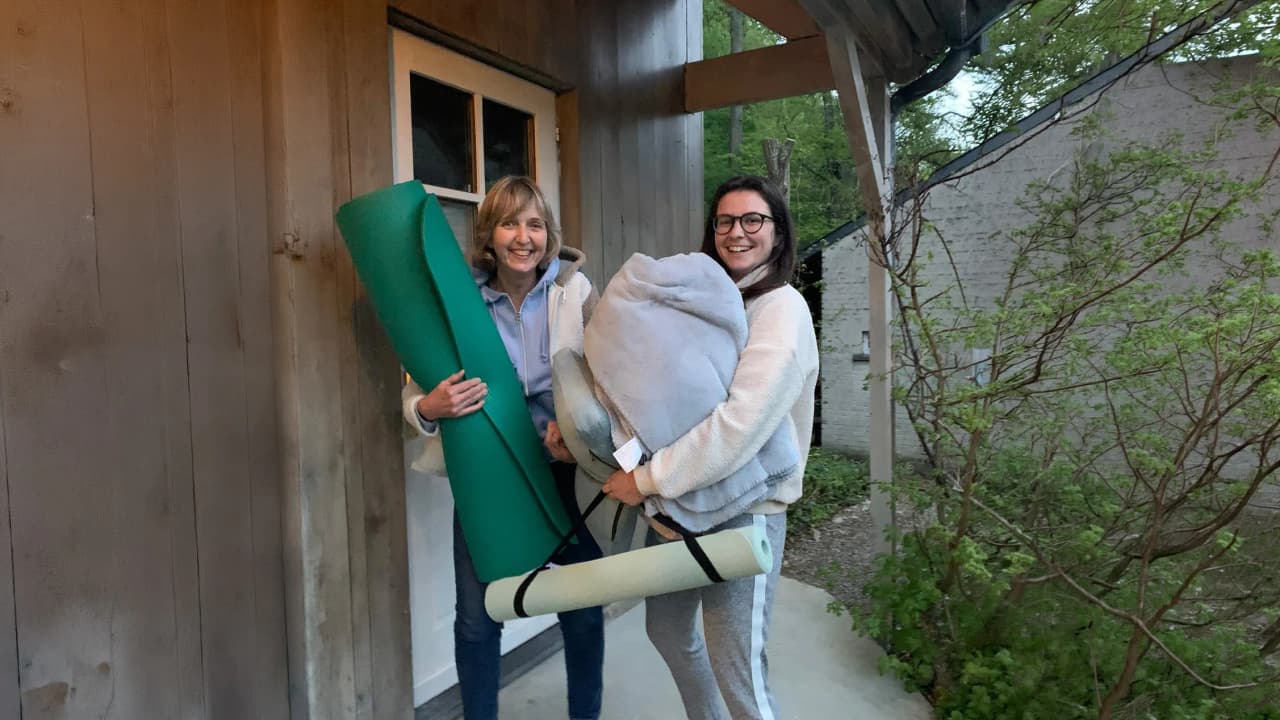 Two smiling women carrying yoga mats and blankets, ready for a nature retreat.