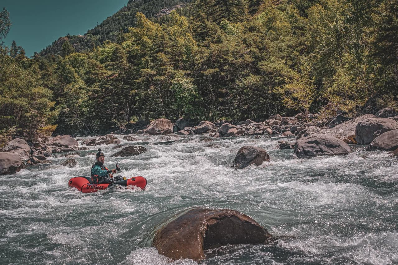 A packraft adventurer navigates a whitewater river, surrounded by cliffs and alpine scenery.