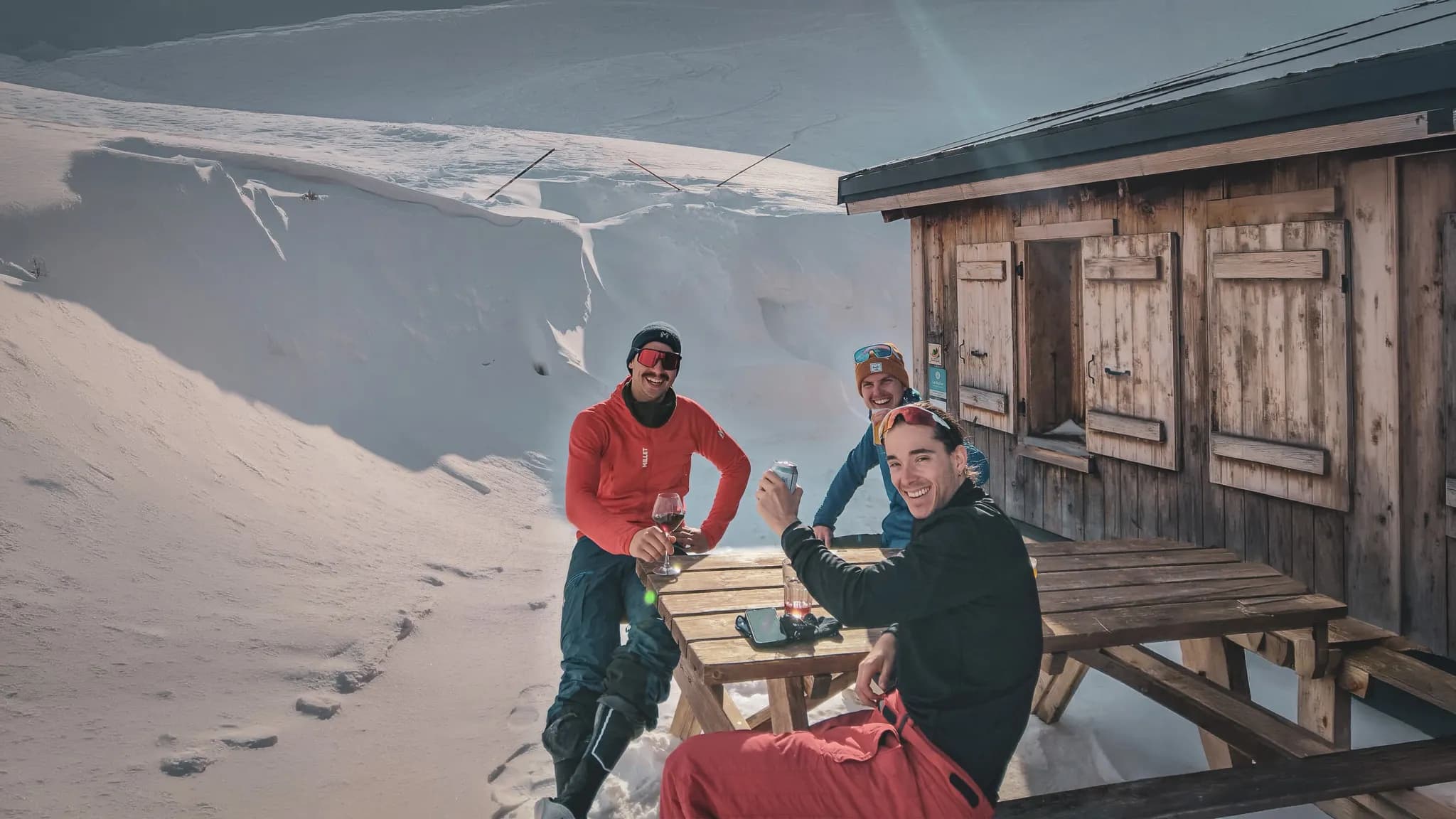 Three friends enjoy the sun and a drink in front of a mountain chalet, surrounded by snow.