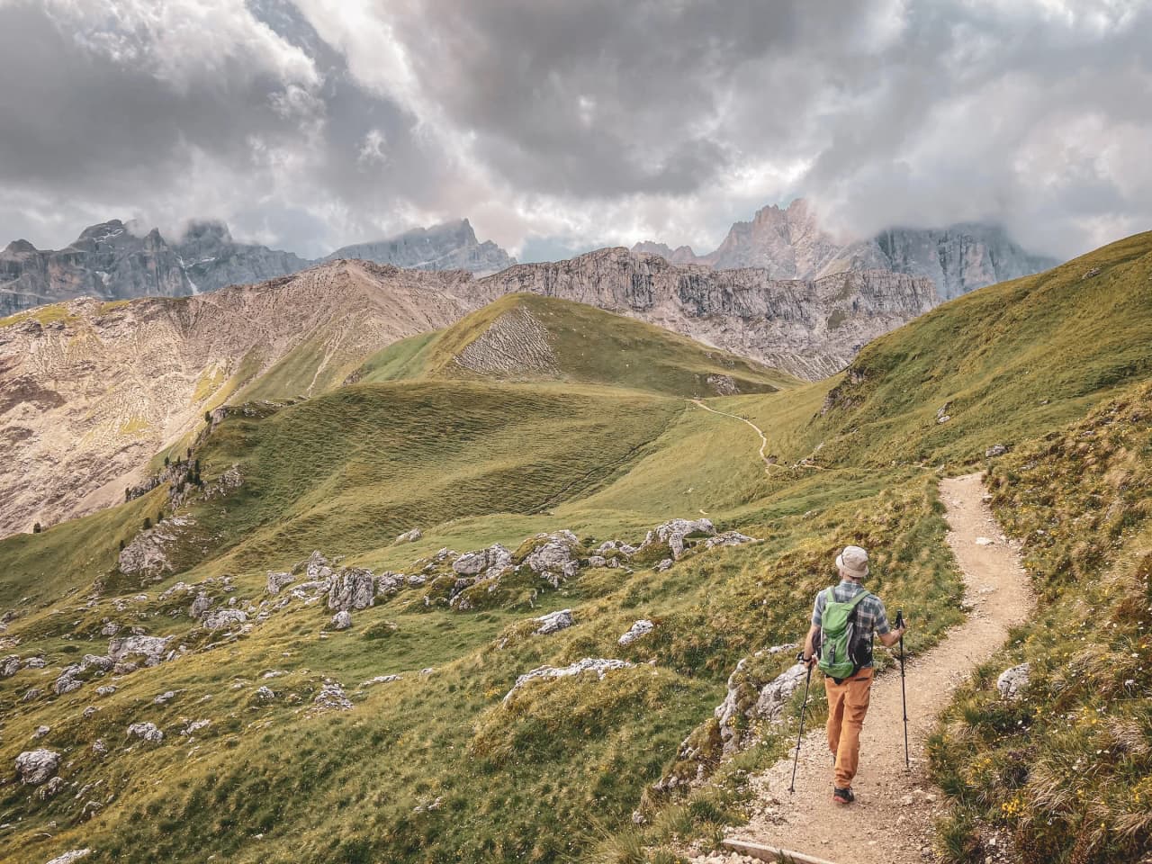 A hiker makes his way along a winding path through a mountainous landscape. Green hills and scattered boulders surround the path, while majestic, cloud-covered peaks