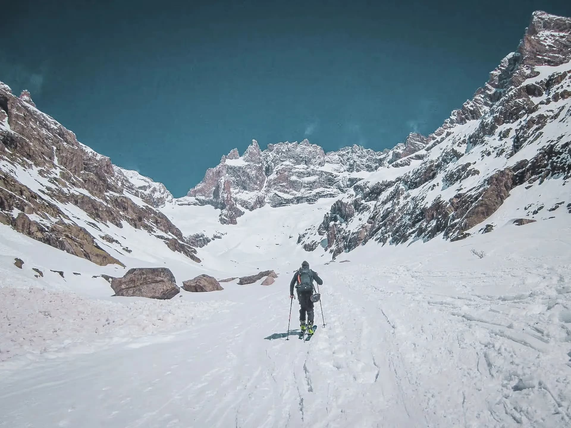 A lone skier advancing through a snow-covered mountain landscape, under a brilliant blue sky.