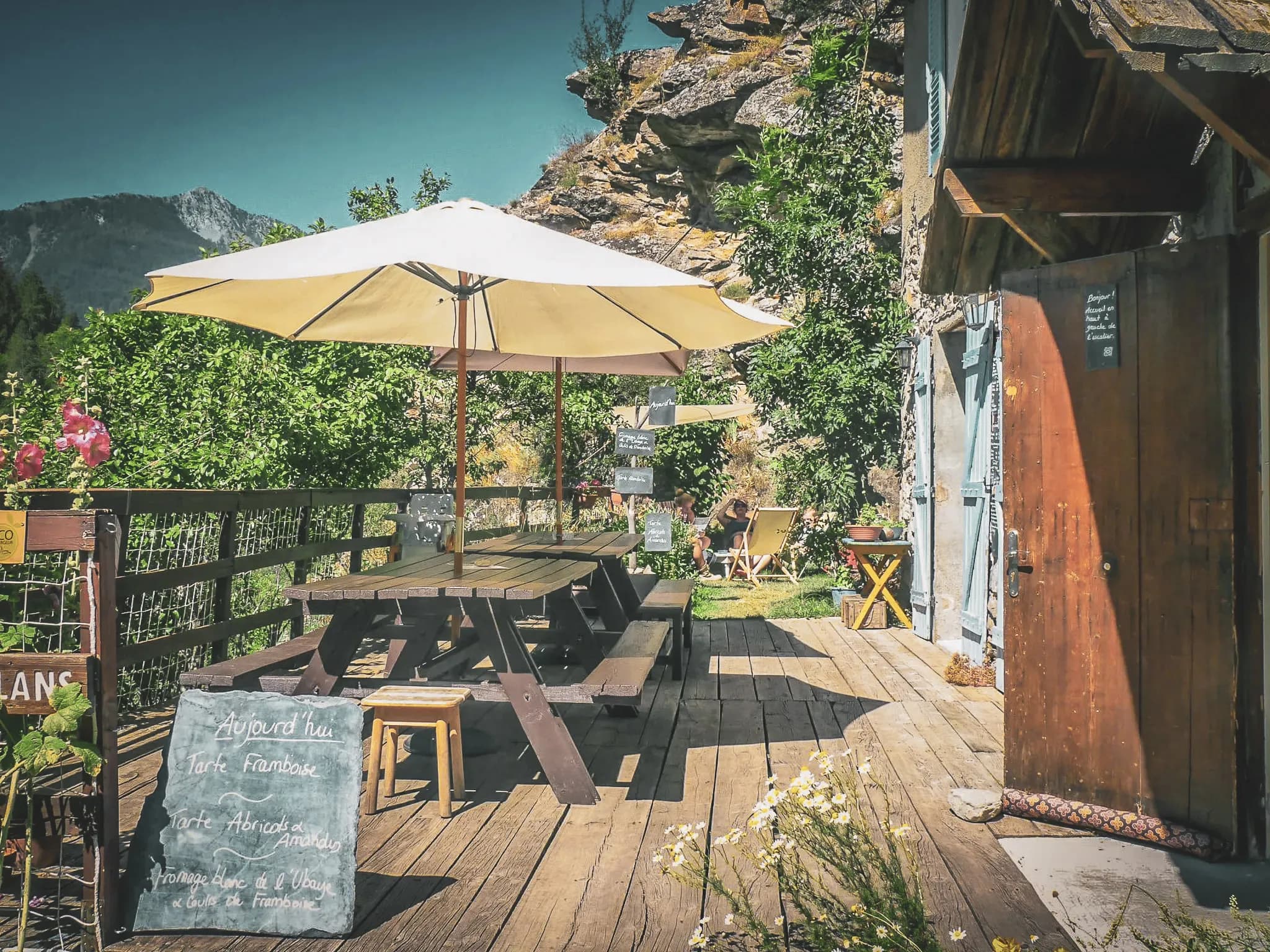 Sunny terrace with parasol, wooden panels, flowers and mountains in the background, peaceful atmosphere.