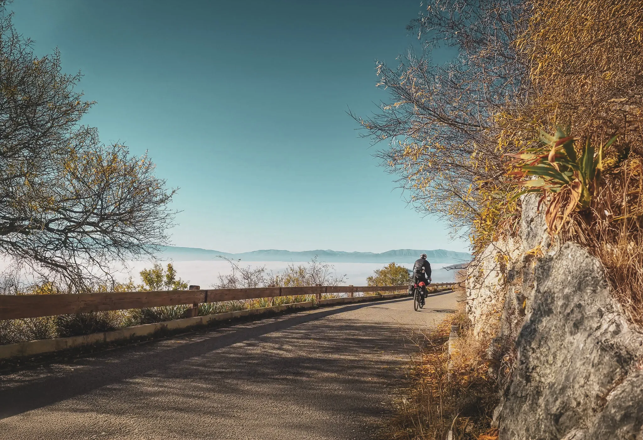Un cycliste pédale le long d'une route sinueuse d'Italie, surplombant une vallée embrumée. Des arbres aux feuilles dorées bordent le chemin, tandis qu'une barrière en bois délimite la route. En arrière-plan
