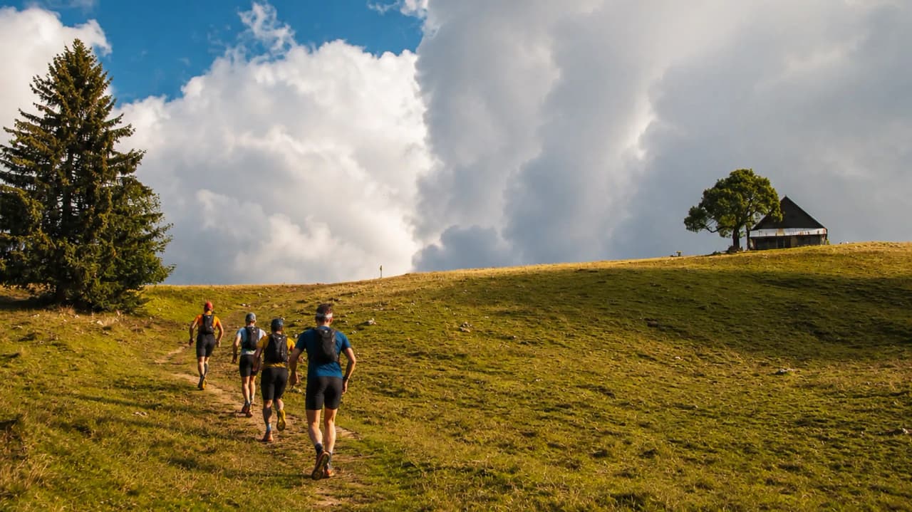 Groupe de randonneurs sur un sentier verdoyant, face à des nuages et une maison en montagne.