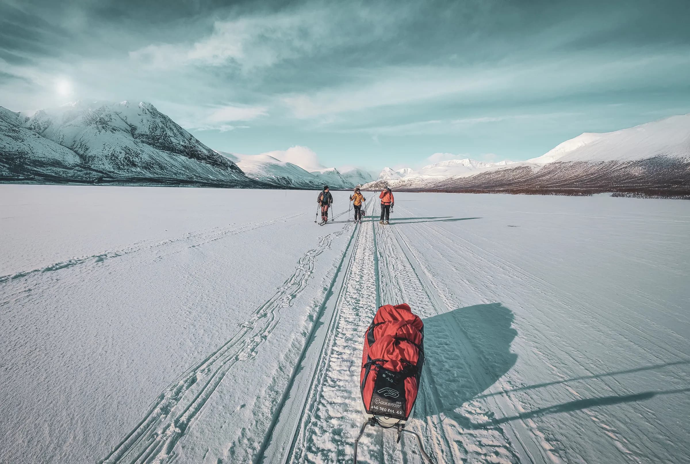An expedition group snowshoes their way across a frozen lake surrounded by snow-capped mountains.