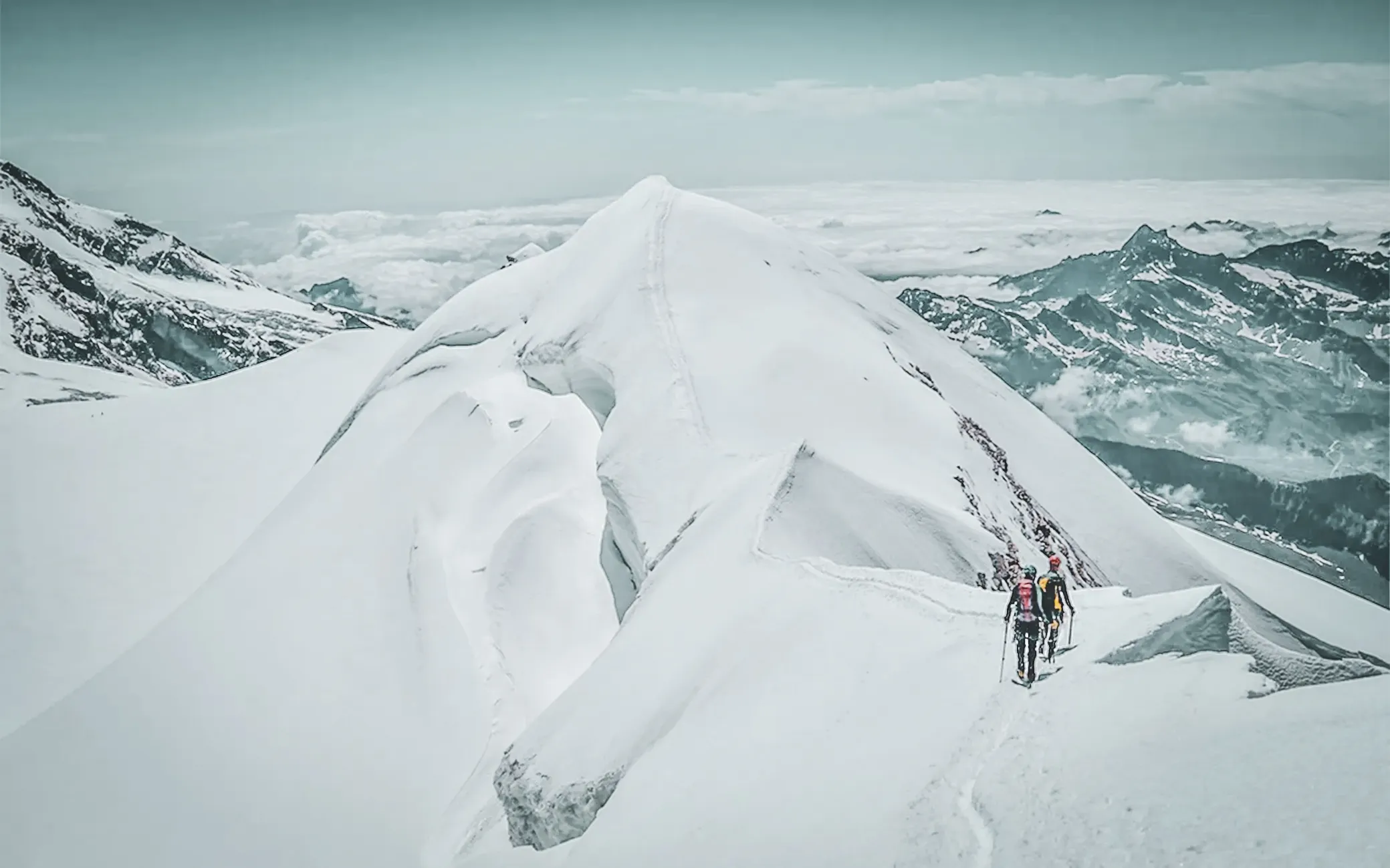 Hikers on a snow-capped peak, overlooking majestic Alpine scenery.