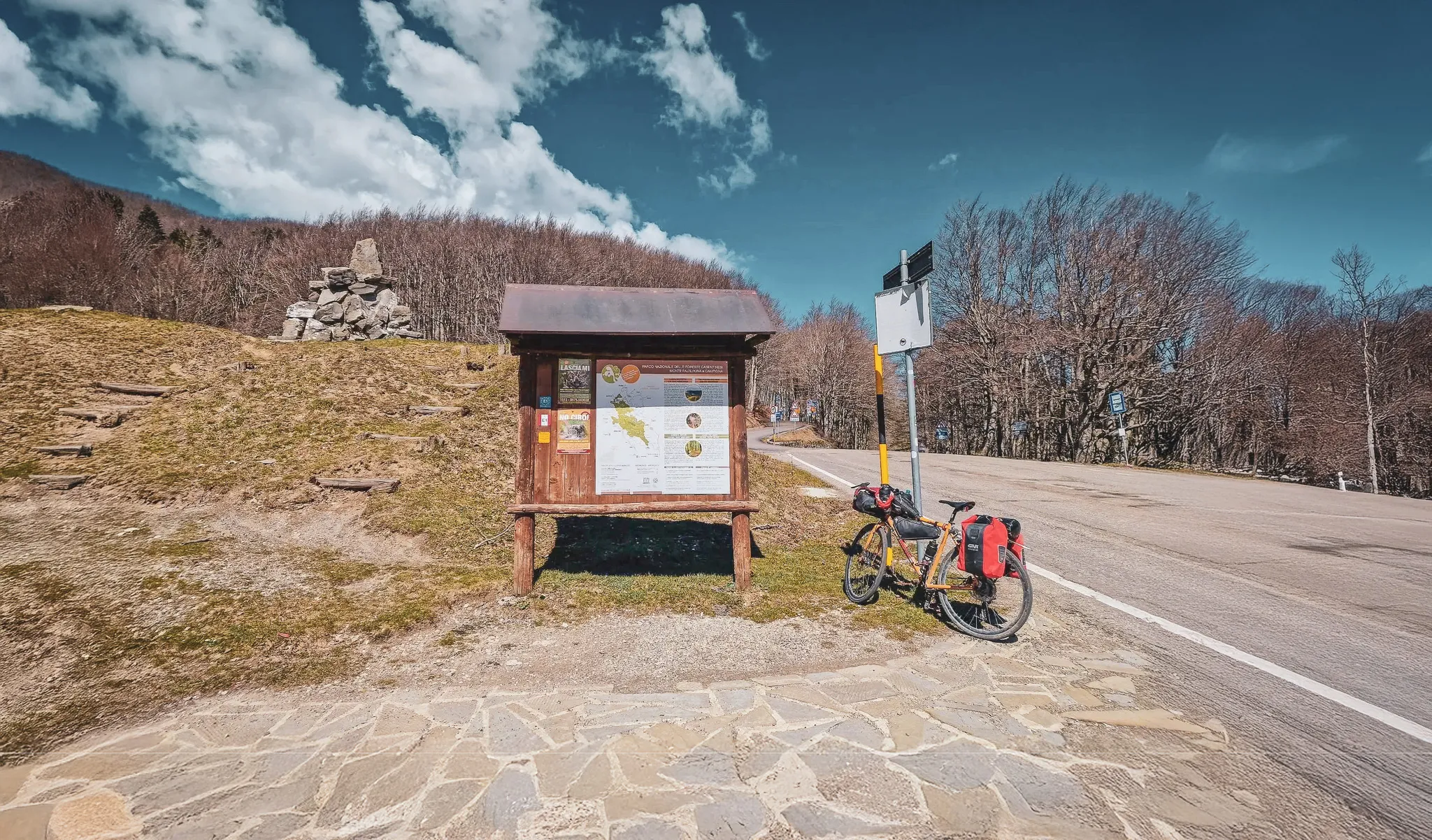 Un panneau d'information en bois se trouve sur un terrain légèrement en pente, entouré de prairies vertes et de quelques arbres dénudés en arrière-plan. À côté du panneau, un vélo avec des sacoches est stationné.