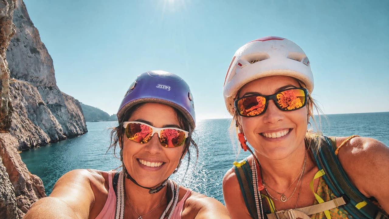 Two smiling female climbers taking a selfie, helmets on their heads, with the sparkling Mediterranean in the background.