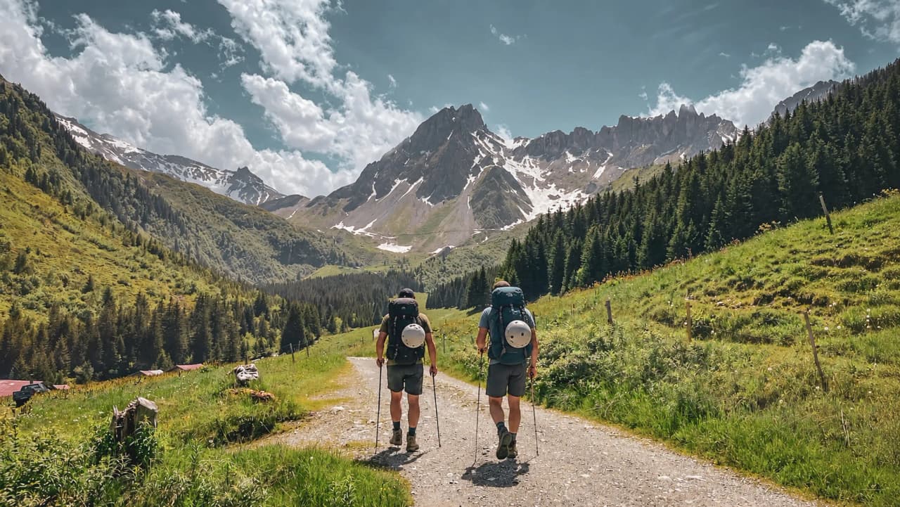 Two hikers walk along a path bordered by greenery, with majestic mountains in the background.