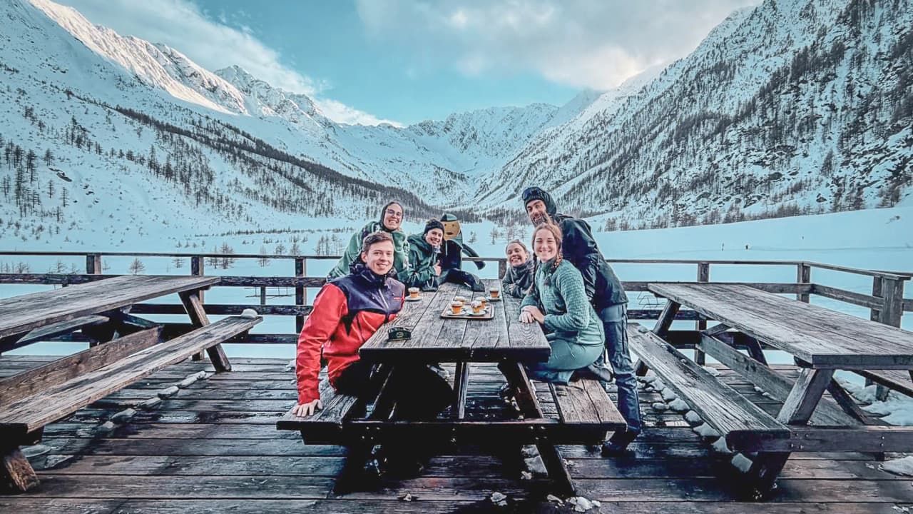 A group of six smiling people on a wooden terrace with snow-capped mountains in the background.