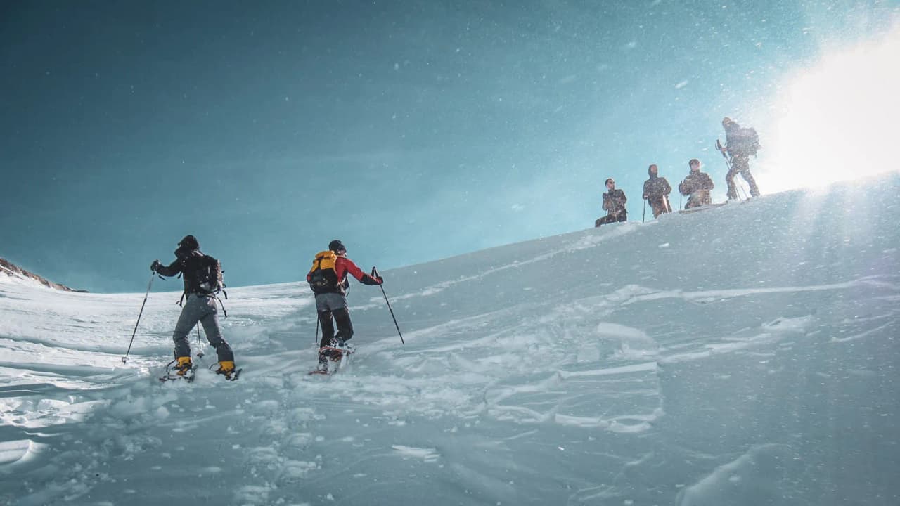 Two people climbing a snow-covered slope, with a group in the background.